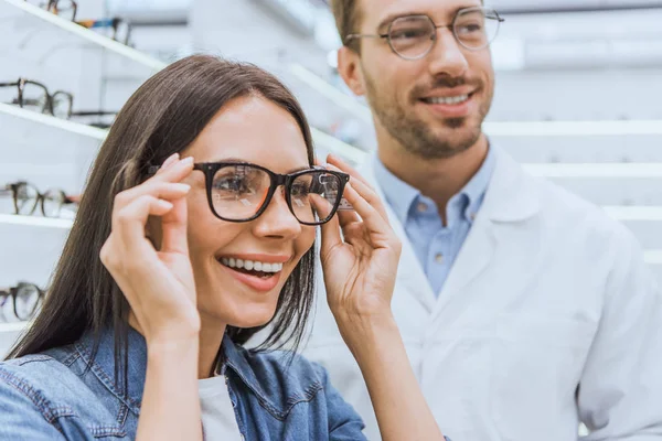 Portrait of smiling woman choosing eyeglasses while male optometrist standing near in optics — Stock Photo