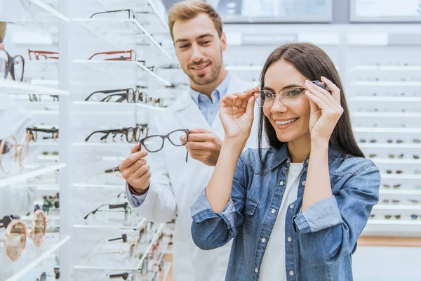 Glückliche Frau wählt Brille, während männlicher Augenarzt in der Nähe mit einer anderen Brille in Optik steht — Stockfoto