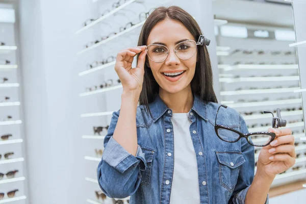 Joyeuse jeune femme choisissant des lunettes et regardant la caméra dans la boutique ophtalmique — Photo de stock
