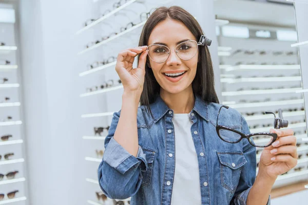 Alegre joven mujer la elección de gafas y mirando a la cámara en la tienda oftálmica - foto de stock