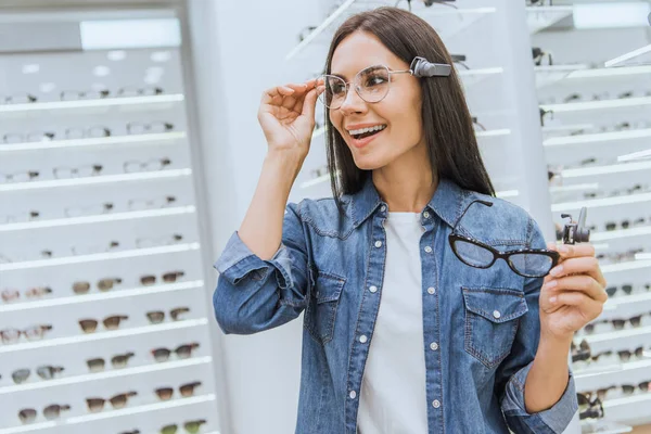 Mujer atractiva sonriente eligiendo gafas en óptica - foto de stock