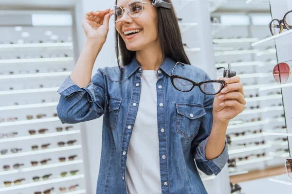 Selective focus of happy young woman choosing eyeglasses in ophthalmic shop — Stock Photo