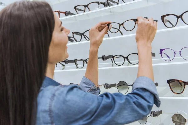 Боковой вид привлекательной молодой женщины, снимающей очки с полок в оптике — стоковое фото