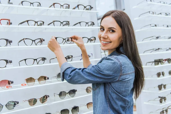 Hermosa joven mujer tomando gafas de los estantes en la óptica — Stock Photo