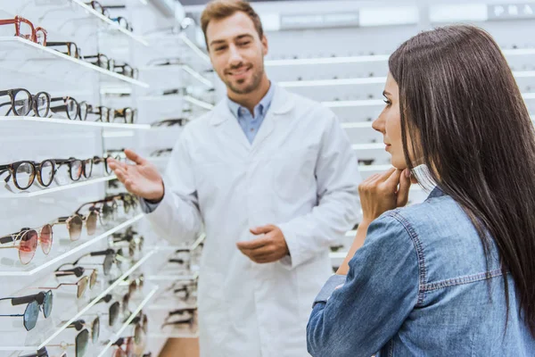 Selective focus of male oculist pointing by hand at shelves with eyeglasses to pensive woman in ophthalmic shop — Stock Photo