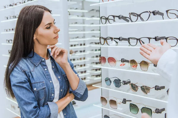 Nachdenkliche Frau blickt auf Regale, während Augenärztin mit der Hand auf Brille in Optik zeigt — Stockfoto