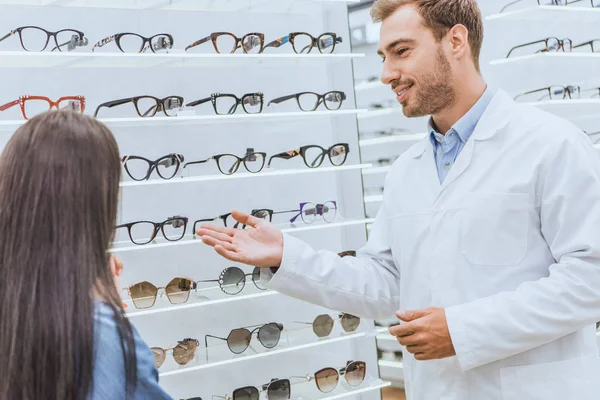 Homem oculista sorrindo apontando à mão em prateleiras com óculos para mulher em óptica — Fotografia de Stock