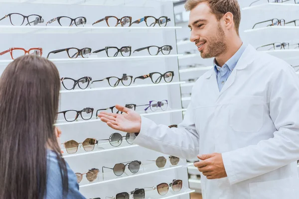 Lächelnder Augenarzt zeigt mit der Hand auf Regale mit Brille auf Frau in Optik — Stock Photo
