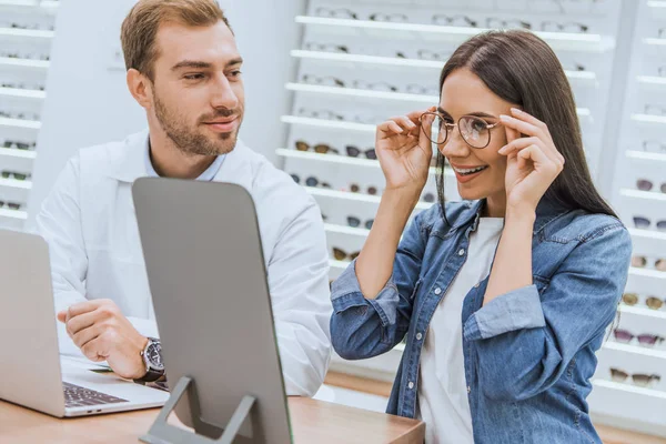 Glückliche Frau, die eine Brille wählt und in den Spiegel schaut, während ein männlicher Augenarzt mit Laptop in der Optik in der Nähe steht — Stockfoto