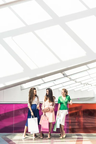 Mujeres jóvenes con estilo sosteniendo bolsas de papel y hablando en el centro comercial - foto de stock