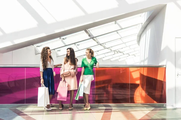 Vista completa de las chicas elegantes sosteniendo bolsas de papel y hablando en el centro comercial - foto de stock