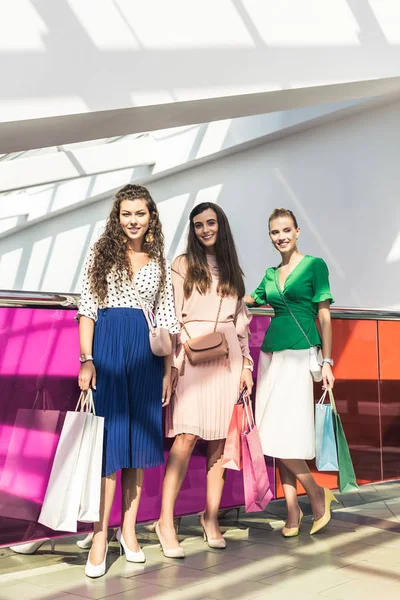 Hermosas chicas de moda sosteniendo bolsas de papel y sonriendo a la cámara mientras están de pie juntos en el centro comercial - foto de stock