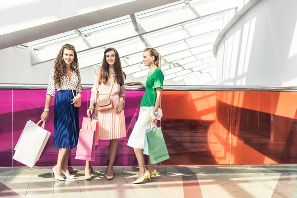 Beautiful stylish girls holding paper bags and smiling at camera in shopping mall — Stock Photo