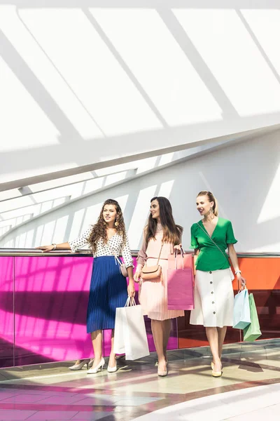 Smiling fashionable girls holding paper bags and looking away at shopping mall — Stock Photo