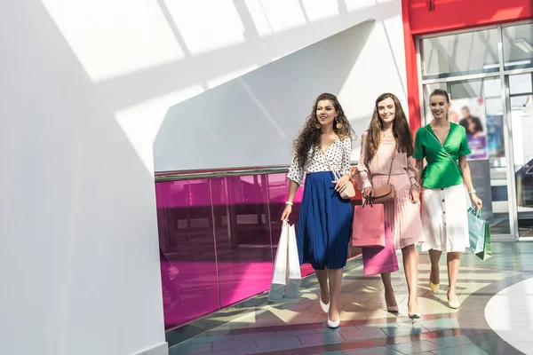 Стильные улыбающиеся молодые женщины, держащие бумажные пакеты и гуляющие вместе в торговом центре — стоковое фото