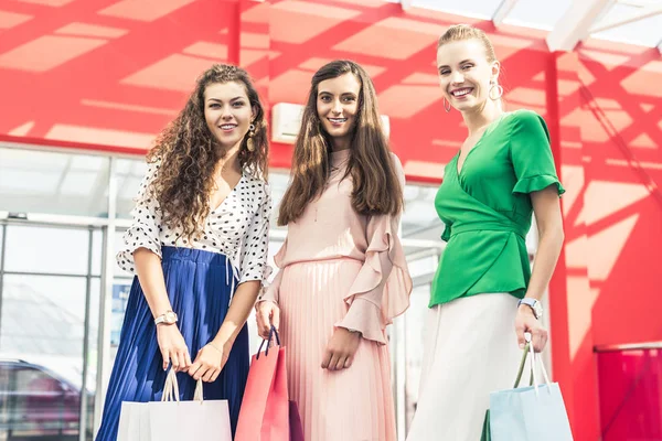 Low angle view of beautiful stylish young women holding paper bags and smiling at camera in shopping mall — Stock Photo
