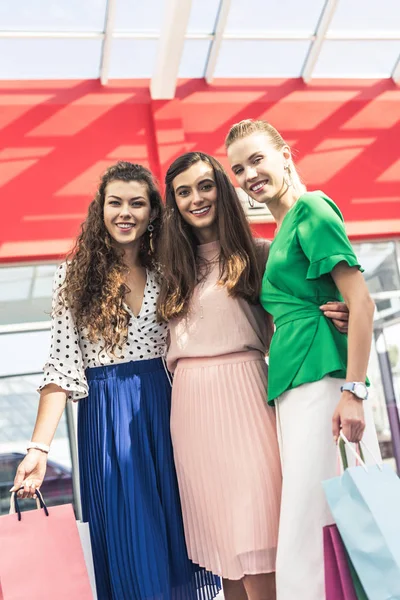 Belas mulheres jovens elegantes segurando sacos de papel e sorrindo para a câmera no shopping — Fotografia de Stock