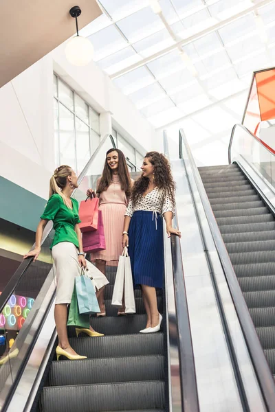 Baixo ângulo de visão de belas mulheres jovens sorridentes segurando sacos de papel na escada rolante no shopping — Fotografia de Stock