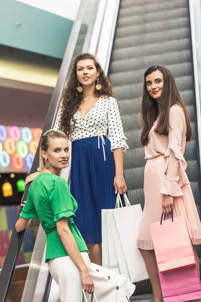 Красиві усміхнені молоді жінки з паперовими мішками, що стоять на ескалаторі в торговому центрі — стокове фото