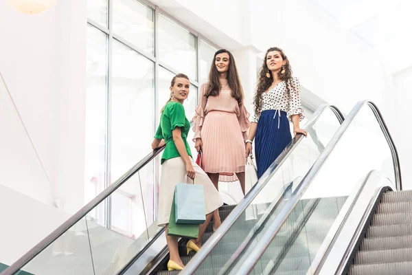 Vista de ángulo bajo de las mujeres jóvenes con estilo sonriente sosteniendo bolsas de compras en escaleras mecánicas - foto de stock