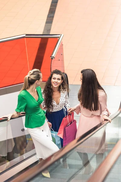 Vue grand angle des filles élégantes souriantes tenant des sacs en papier et parlant sur l'escalator dans le centre commercial — Photo de stock