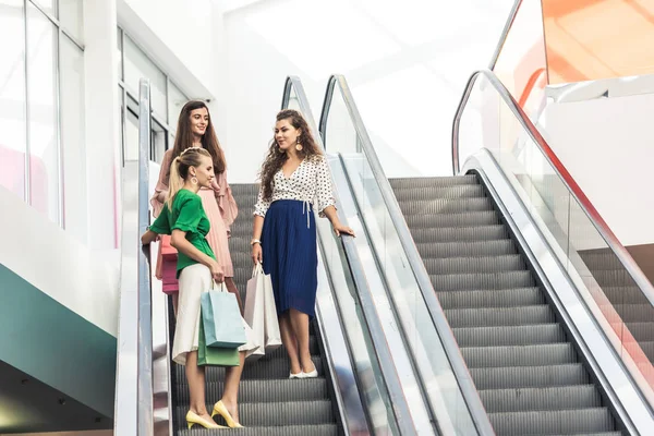 Vista de ángulo bajo de chicas sonrientes con estilo sosteniendo bolsas de compras en escaleras mecánicas - foto de stock