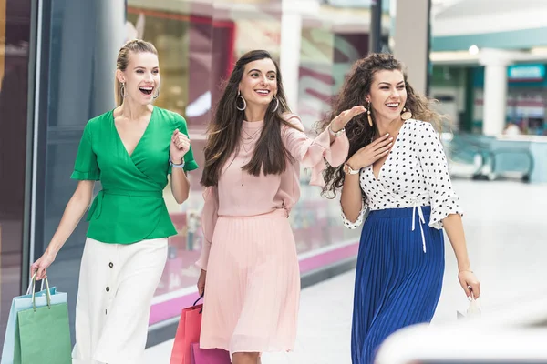 Excited young women holding paper bags and looking away while walking together in shopping mall — Stock Photo