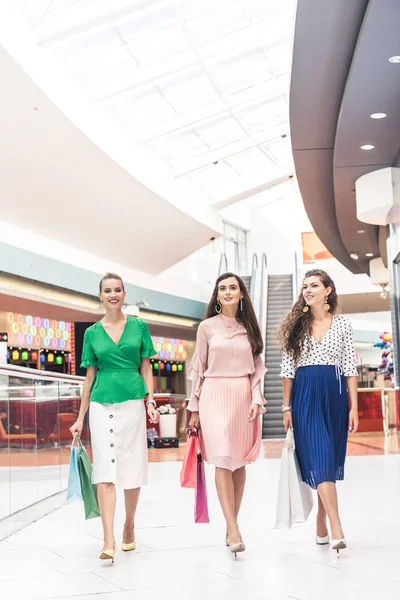 Élégantes jeunes femmes souriantes tenant des sacs en papier et marchant ensemble dans un centre commercial — Photo de stock