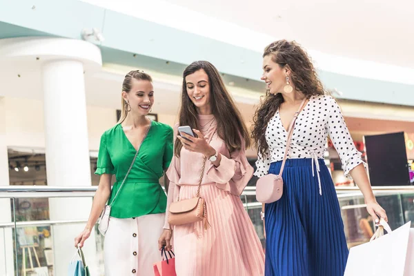 Beautiful smiling girls holding paper bags and using smartphone in shopping mall — Stock Photo
