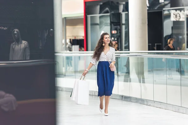 Вид в полный рост улыбающейся стильной молодой женщины, держащей в руках сумки с покупками и ходящей в грязи — стоковое фото