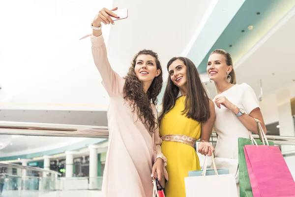 Vue à angle bas de filles élégantes heureuses tenant des sacs en papier et prenant selfie avec smartphone dans le centre commercial — Photo de stock