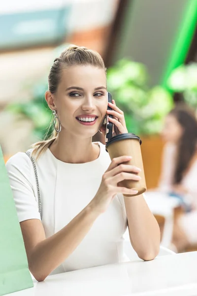 Beautiful smiling girl holding disposable coffee cup and talking by smartphone while sitting in cafe at shopping mall — Stock Photo