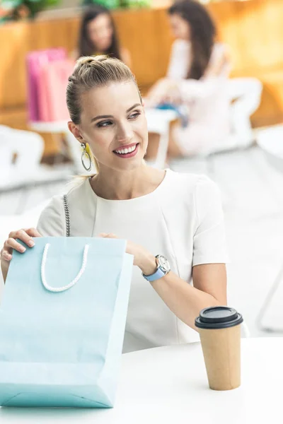 Hermosa mujer joven sonriente sosteniendo bolsa de papel y mirando hacia otro lado en el café en el centro comercial - foto de stock