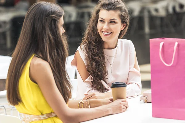 Belas jovens mulheres sorrindo umas às outras e falando enquanto se sentam à mesa no café no shopping — Fotografia de Stock