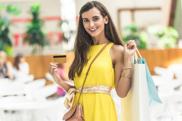 Bella giovane donna sorridente in possesso di sacchetti di carta e carta di credito nel centro commerciale — Foto stock