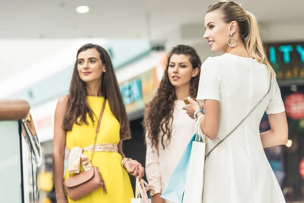 Hermosas mujeres jóvenes de moda sosteniendo bolsas de papel y mirando hacia el centro comercial - foto de stock