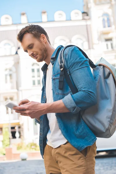 Vista inferior del joven guapo con mochila caminando por la calle y utilizando el teléfono inteligente - foto de stock