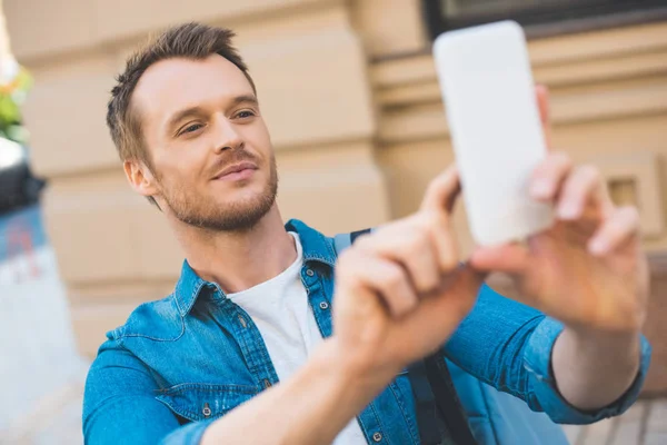 Close-up portrait of handsome young tourist with backpack taking photo with smartphone on street — Stock Photo