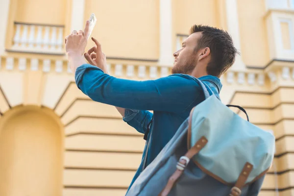 Handsome young tourist with backpack taking photo with smartphone on street — Stock Photo