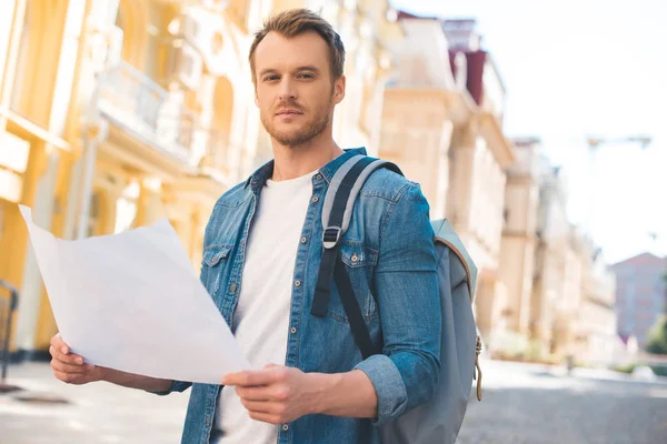 Handsome young tourist with backpack and map looking at camera on street — Stock Photo