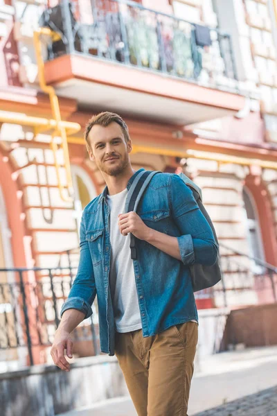 Sonriente joven viajero con mochila caminando por la calle y mirando a la cámara - foto de stock