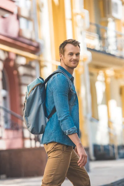 Jovem viajante atraente com mochila andando pela rua e olhando para a câmera — Fotografia de Stock