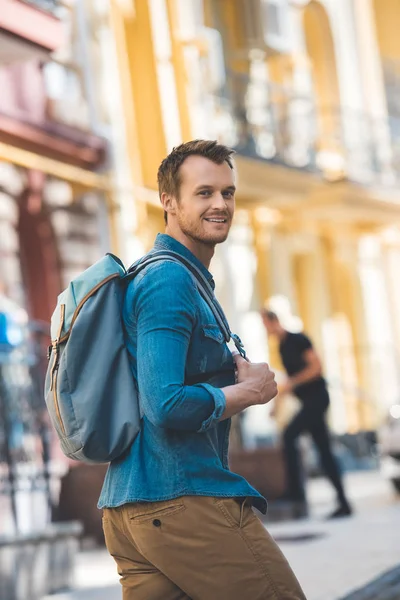 Счастливый молодой человек с рюкзаком ходит по улице и смотрит в камеру — стоковое фото