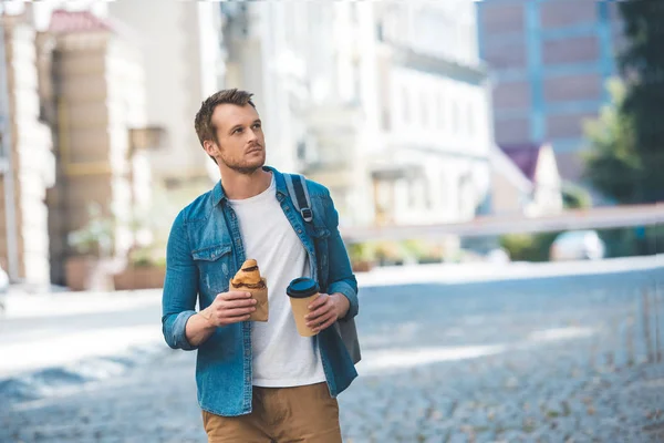 Handsome young man with backpack, coffee to go and croissant walking by street and looking away — Stock Photo