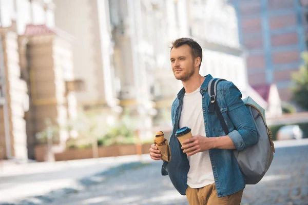 Handsome young man with backpack, coffee to go and croissant walking by street and looking at camera — Stock Photo