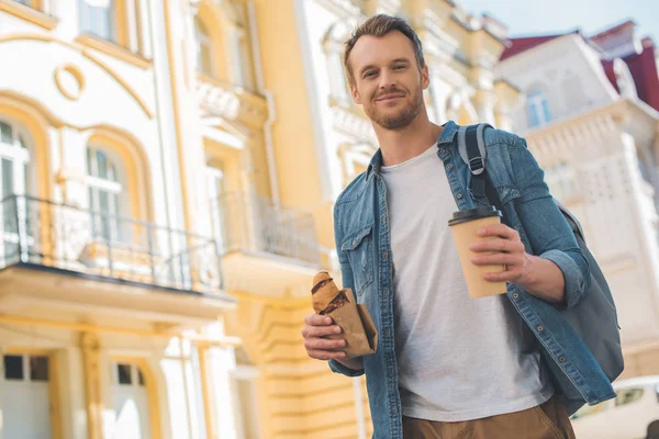 Vue du bas de beau jeune homme avec sac à dos, café à emporter et croissant marchant dans la rue et regardant la caméra — Photo de stock