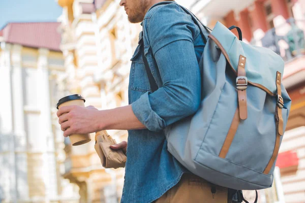 Cropped shot of young man with backpack, coffee to go and croissant walking by street — Stock Photo