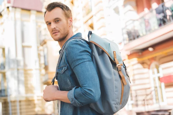Handsome young man with backpack walking by street and looking at camera — Stock Photo