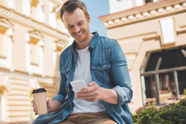 Heureux jeune homme avec café pour aller à l'aide d'un smartphone sur la rue — Photo de stock