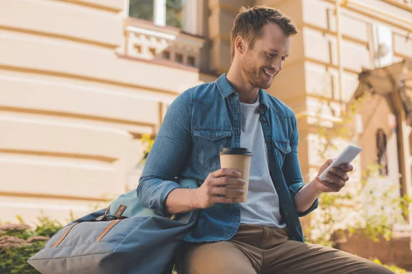 Bel giovane uomo con tazza di carta di caffè utilizzando smartphone sulla strada — Foto stock